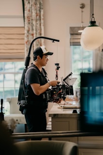 A person wearing a cap and a harness operates a professional camera rig inside a well-lit room. The room has modern decor with patterned curtains and a large pendant light hanging from the ceiling. The person is focused on the camera, which has multiple attachments and cables.
