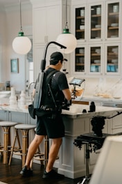 A person wearing a black outfit and a cap operates a camera stabilizer rig in a kitchen setting. The kitchen features white cabinetry, two spherical pendant lights, a marble countertop, and several bar stools. The person appears to be filming or setting up for a shoot.