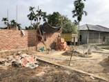 A construction site with partially built brick walls, piles of bricks, sand, and cement scattered around. There are several construction tools including shovels and a cement mixer. A tarp is loosely draped over some scaffolding, and palm trees are in the background with a truck parked nearby.