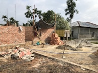 A construction site with partially built brick walls, piles of bricks, sand, and cement scattered around. There are several construction tools including shovels and a cement mixer. A tarp is loosely draped over some scaffolding, and palm trees are in the background with a truck parked nearby.