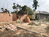 A construction site with partially built brick walls, piles of bricks, sand, and cement scattered around. There are several construction tools including shovels and a cement mixer. A tarp is loosely draped over some scaffolding, and palm trees are in the background with a truck parked nearby.