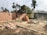 A construction site with partially built brick walls, piles of bricks, sand, and cement scattered around. There are several construction tools including shovels and a cement mixer. A tarp is loosely draped over some scaffolding, and palm trees are in the background with a truck parked nearby.