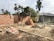 A construction site with partially built brick walls, piles of bricks, sand, and cement scattered around. There are several construction tools including shovels and a cement mixer. A tarp is loosely draped over some scaffolding, and palm trees are in the background with a truck parked nearby.