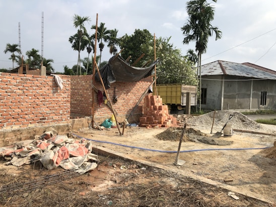 A construction site with partially built brick walls, piles of bricks, sand, and cement scattered around. There are several construction tools including shovels and a cement mixer. A tarp is loosely draped over some scaffolding, and palm trees are in the background with a truck parked nearby.