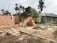 A construction site with partially built brick walls, piles of bricks, sand, and cement scattered around. There are several construction tools including shovels and a cement mixer. A tarp is loosely draped over some scaffolding, and palm trees are in the background with a truck parked nearby.