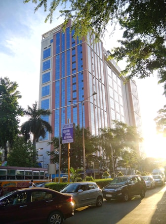 A tall, modern building with a facade of blue-tinted glass panels and reddish-brown accents stands alongside a busy street. Numerous vehicles, including cars and a bus, are seen on the road. There are trees and greenery in the surrounding area, with sunlight streaming through the leaves, casting shadows on the ground. A sign for a public school is displayed near the building.