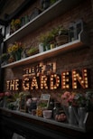 A rustic brick wall adorned with shelves holding various potted plants, vibrant flowers, and decorative lanterns. Below the shelves, a marquee light sign spells out 'THE GARDEN' in large, illuminated letters. Assorted garden-themed decor, such as watering cans and jars, are neatly arranged on the lower shelf.