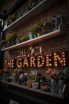 A rustic brick wall adorned with shelves holding various potted plants, vibrant flowers, and decorative lanterns. Below the shelves, a marquee light sign spells out 'THE GARDEN' in large, illuminated letters. Assorted garden-themed decor, such as watering cans and jars, are neatly arranged on the lower shelf.