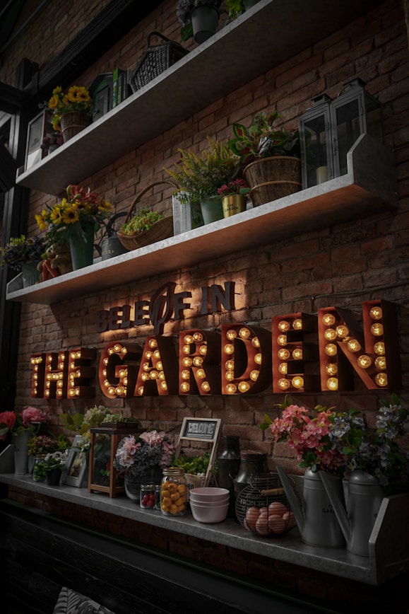 A rustic brick wall adorned with shelves holding various potted plants, vibrant flowers, and decorative lanterns. Below the shelves, a marquee light sign spells out 'THE GARDEN' in large, illuminated letters. Assorted garden-themed decor, such as watering cans and jars, are neatly arranged on the lower shelf.