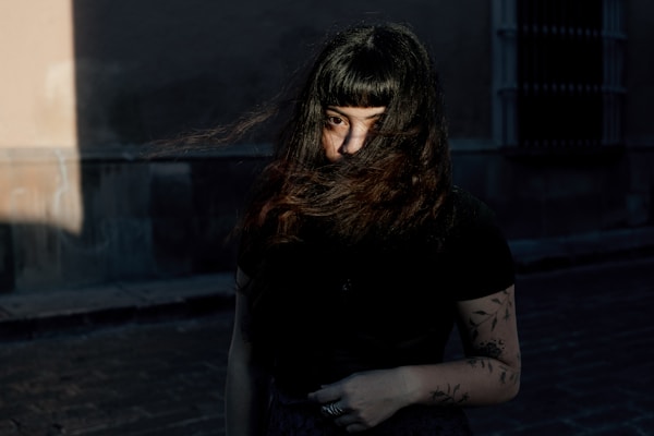 A person with long, dark hair partially obscuring their face stands in a dimly lit urban setting. Tattoos are visible on the arm, and there is a mix of natural and artificial lighting, casting shadows and highlighting parts of the scene.