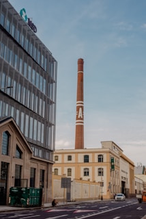An urban street scene featuring industrial buildings and a tall brick smokestack with a large 'A' painted on it. The foreground includes a modern building with large windows, while the background shows older industrial architecture. A lone pedestrian is crossing the street, and parked vehicles are visible.
