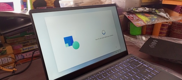 A laptop with a visible startup screen displaying text and a loading icon. Surrounding the laptop are various items such as board games, colorful bangles, and plastic containers. The keyboard is lit with a soft glow, and the overall setting appears to be cluttered, possibly a personal or creative workspace.