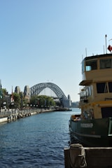 A large ferry named 'Friendship' is docked along a waterfront with a row of lamp posts and flags lining the dock. In the background, there is a prominent steel arch bridge spanning a body of water. The sky is clear and blue, contributing to a serene and pleasant atmosphere.