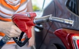 A person is holding a red gasoline pump nozzle next to a vehicle, with a blurred background. The focus is on the nozzle and hand, indicating a refueling activity.