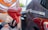 A person is holding a red gasoline pump nozzle next to a vehicle, with a blurred background. The focus is on the nozzle and hand, indicating a refueling activity.