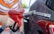 A person is holding a red gasoline pump nozzle next to a vehicle, with a blurred background. The focus is on the nozzle and hand, indicating a refueling activity.