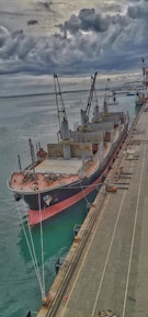 A large cargo ship is docked at a port with multiple cranes on its deck. The scene is overcast, with dark clouds looming in the sky, adding a dramatic effect. The waters of the port are calm, reflecting the grays of the sky. The dock is seen alongside the ship with various equipment and materials.