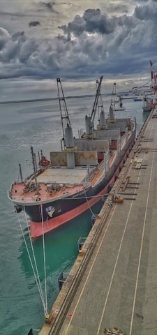 A large cargo ship is docked at a port with multiple cranes on its deck. The scene is overcast, with dark clouds looming in the sky, adding a dramatic effect. The waters of the port are calm, reflecting the grays of the sky. The dock is seen alongside the ship with various equipment and materials.