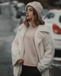 A person stands outdoors wearing a cozy cream-colored coat, a beige knitted hat, and dark pants. The background features a blurry street with parked cars and a slightly overcast atmosphere.