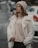 A person stands outdoors wearing a cozy cream-colored coat, a beige knitted hat, and dark pants. The background features a blurry street with parked cars and a slightly overcast atmosphere.