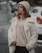 A person stands outdoors wearing a cozy cream-colored coat, a beige knitted hat, and dark pants. The background features a blurry street with parked cars and a slightly overcast atmosphere.