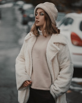 A person stands outdoors wearing a cozy cream-colored coat, a beige knitted hat, and dark pants. The background features a blurry street with parked cars and a slightly overcast atmosphere.