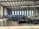 A large industrial warehouse with a high ceiling and several tall windows allowing natural light to enter. There are stacks of blue plastic bags or containers lined up against the back wall. Black tarps or materials are spread across the center of the concrete floor. Various equipment and boxes are organized along the right side of the space.