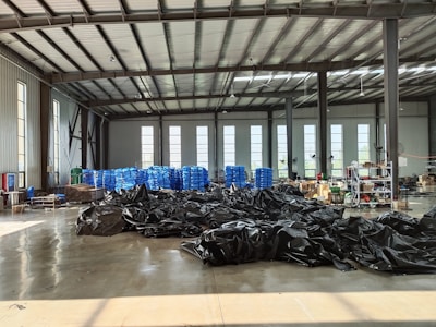 A large industrial warehouse with a high ceiling and several tall windows allowing natural light to enter. There are stacks of blue plastic bags or containers lined up against the back wall. Black tarps or materials are spread across the center of the concrete floor. Various equipment and boxes are organized along the right side of the space.