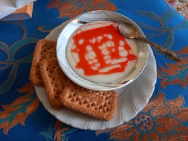 A bowl of creamy white dessert with red syrup is placed on a white plate, accompanied by four rectangular cookies. A spoon rests on the edge of the bowl. The setup is on a vibrant blue cloth with decorative orange and teal floral patterns.