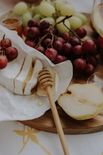 A wooden platter contains fresh red and green grapes, sliced brie cheese, a honey dipper with drips of honey, and a halved pear. The setting appears to be part of a rustic picnic or cheese and fruit board setup.