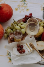 A wooden platter holds an assortment of fruits including red and green grapes, along with a glass of white wine nearby. There is a wedge of brie cheese topped with red grapes, a honey dipper, jars of preserves, and a sliced pear on a white tablecloth with yellow floral patterns. Two pumpkins can be seen on the left side of the image.
