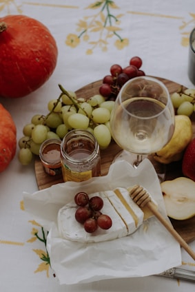 A wooden platter holds an assortment of fruits including red and green grapes, along with a glass of white wine nearby. There is a wedge of brie cheese topped with red grapes, a honey dipper, jars of preserves, and a sliced pear on a white tablecloth with yellow floral patterns. Two pumpkins can be seen on the left side of the image.