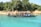 A long black boat with a canopy is floating on clear turquoise water. Several people are seated on the boat, wearing life jackets. The boat is close to a shore, where there are dense green trees and some brown and beige rocks.