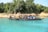 A long black boat with a canopy is floating on clear turquoise water. Several people are seated on the boat, wearing life jackets. The boat is close to a shore, where there are dense green trees and some brown and beige rocks.