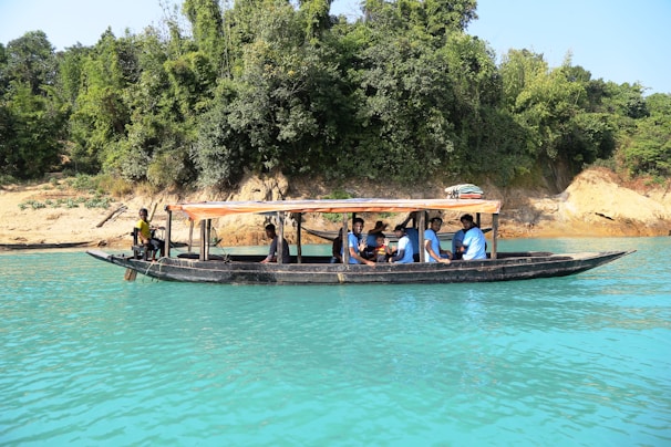 A long black boat with a canopy is floating on clear turquoise water. Several people are seated on the boat, wearing life jackets. The boat is close to a shore, where there are dense green trees and some brown and beige rocks.