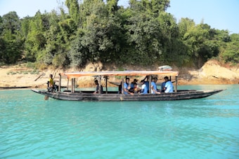 A long black boat with a canopy is floating on clear turquoise water. Several people are seated on the boat, wearing life jackets. The boat is close to a shore, where there are dense green trees and some brown and beige rocks.