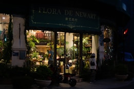 A shop with a lush display of plants visible through large windows, illuminated from inside. The exterior features greenery and a sign reading 'Flora De Newark' with additional text in another language. A person stands outside, looking into the shop, with a bicycle parked nearby. The scene is set in the evening, adding a warm, inviting atmosphere.