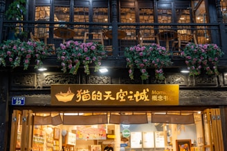 A charming café facade features a wooden sign with yellow and white lettering, surrounded by decorative wooden panels. The balcony above is adorned with hanging baskets of pink and white flowers, and the warm interior lighting is visible through the large glass windows.