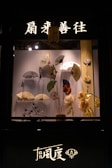 A display window features a collection of decorative hand fans arranged artfully against a backdrop. Above the display, illuminated Chinese characters and a lantern add an authentic touch. There are flowers and other decorative items complementing the fans.