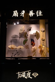A display window features a collection of decorative hand fans arranged artfully against a backdrop. Above the display, illuminated Chinese characters and a lantern add an authentic touch. There are flowers and other decorative items complementing the fans.