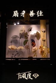 A display window features a collection of decorative hand fans arranged artfully against a backdrop. Above the display, illuminated Chinese characters and a lantern add an authentic touch. There are flowers and other decorative items complementing the fans.