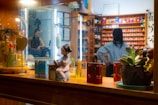 A vibrant storefront window display filled with various decorative items, including a doll dressed in traditional attire, lush green succulents in a decorative planter, colorful cosmetic products, and a small golden figurine. In the background, a warmly-lit shop interior is visible, displaying shelves lined with numerous small, colorful boxes. Reflections of two people can be seen, one standing with hands on hips, suggesting a busy or retail setting.