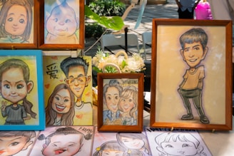 A collection of framed caricature portraits displayed on a table. The caricatures include a variety of people featuring exaggerated facial expressions and distinct hairstyles. The drawings are colorful, with some frames made of wood, and a floral decoration is partly visible on the table. The setting appears to be outdoors, as suggested by the natural light and presence of greenery in the background.