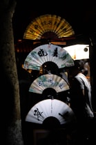A display of decorative paper fans, each with intricate designs and calligraphy, is arranged vertically against a dark background. The fans are illuminated by a soft light source, casting a warm glow. A silhouetted figure stands nearby, adding to the serene ambiance.
