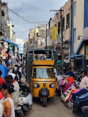 A bustling street market scene with a yellow auto-rickshaw in the center. Various stalls on either side display colorful clothing and accessories. People are interacting and moving around, contributing to a lively atmosphere. Overhead, numerous electrical wires crisscross between buildings, and signs hang from the structures.