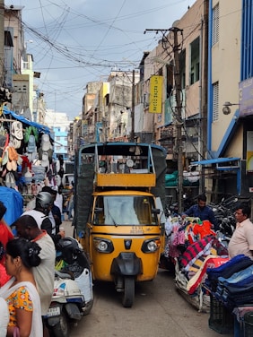 A bustling street market scene with a yellow auto-rickshaw in the center. Various stalls on either side display colorful clothing and accessories. People are interacting and moving around, contributing to a lively atmosphere. Overhead, numerous electrical wires crisscross between buildings, and signs hang from the structures.