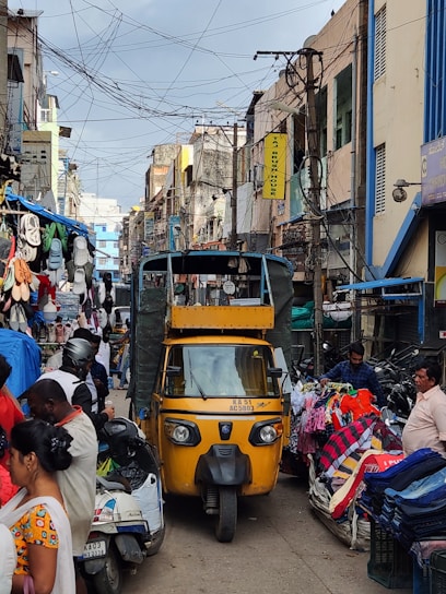 A bustling street market scene with a yellow auto-rickshaw in the center. Various stalls on either side display colorful clothing and accessories. People are interacting and moving around, contributing to a lively atmosphere. Overhead, numerous electrical wires crisscross between buildings, and signs hang from the structures.