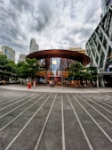 A modern glass-fronted store with a large circular wooden roof located in an urban setting. The Apple logo is prominently displayed above the entrance, which is surrounded by green trees. Skyscrapers and overcast skies are visible in the background. People are entering and exiting the store, contributing to a bustling atmosphere.