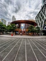 A modern glass-fronted store with a large circular wooden roof located in an urban setting. The Apple logo is prominently displayed above the entrance, which is surrounded by green trees. Skyscrapers and overcast skies are visible in the background. People are entering and exiting the store, contributing to a bustling atmosphere.