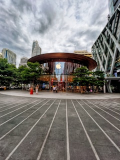 A modern glass-fronted store with a large circular wooden roof located in an urban setting. The Apple logo is prominently displayed above the entrance, which is surrounded by green trees. Skyscrapers and overcast skies are visible in the background. People are entering and exiting the store, contributing to a bustling atmosphere.